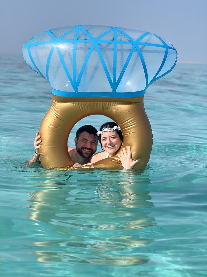 Jessica and Rami peeking through a giant diamond-ring float in turquoise water