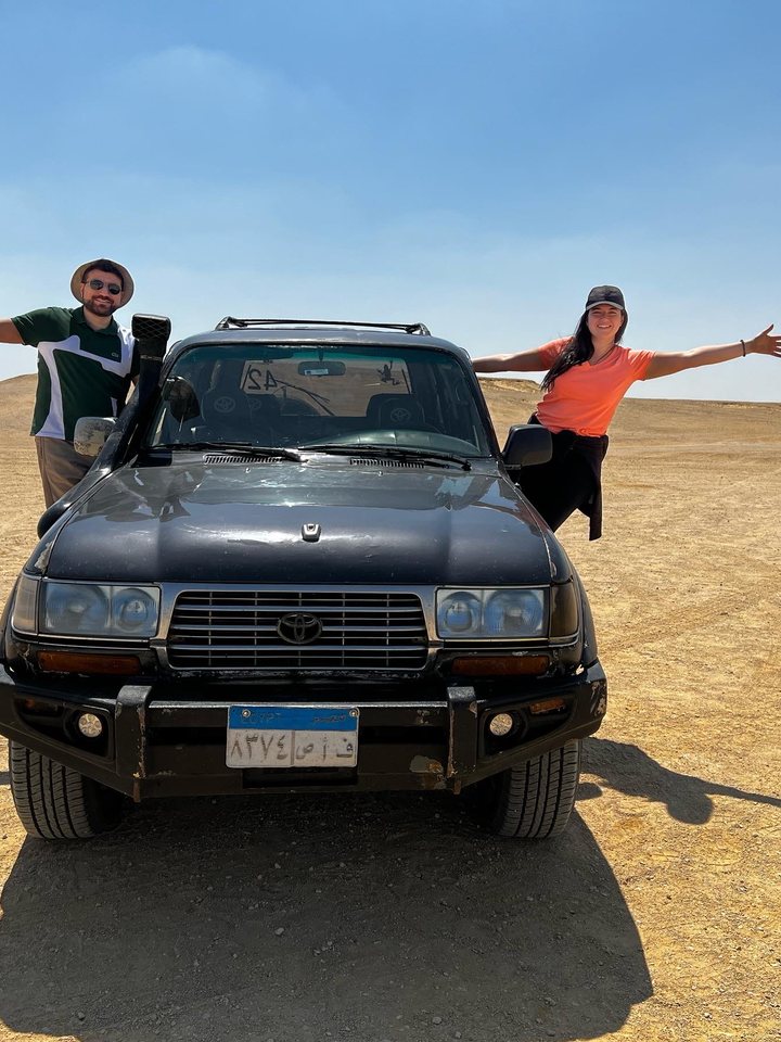 Jessica and Rami with arms outstretched on top of a jeep in the desert