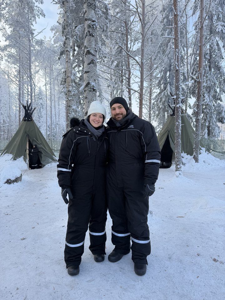 Jessica and Rami in snowsuits in a snowy forest with tipis behind them