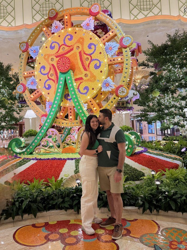 Rami kissing Jessica on the cheek in front of a giant flower Ferris wheel
